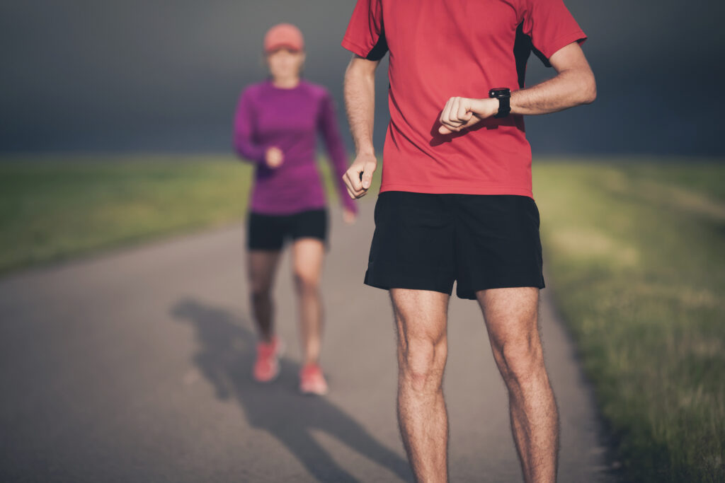 Man and woman runners running on country road in summer sunset. Young training and doing workout outdoors in nature. Checking performance on stopwatch smart watch.